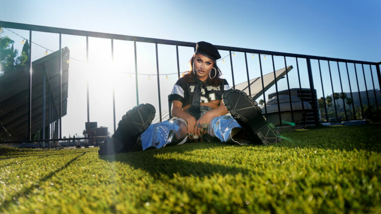 Wide shot of Sasha Colby sitting by rails on grass in jeans and a black beret
