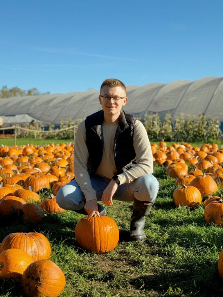 A man crouches down in a field of pumpkins and looks at the camera smiling