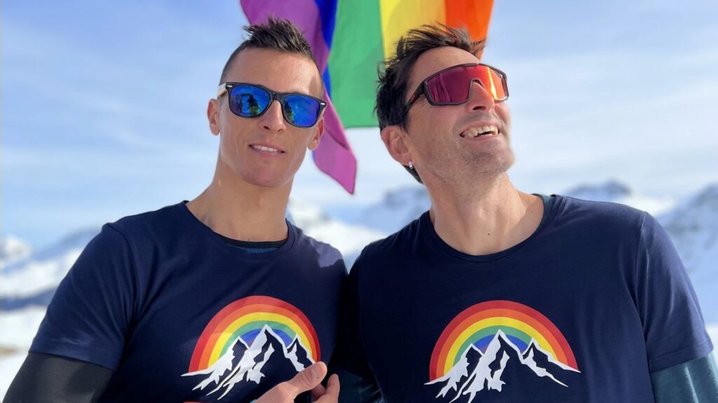 Two men stand on a snowy mountain with a Pride flag behind them