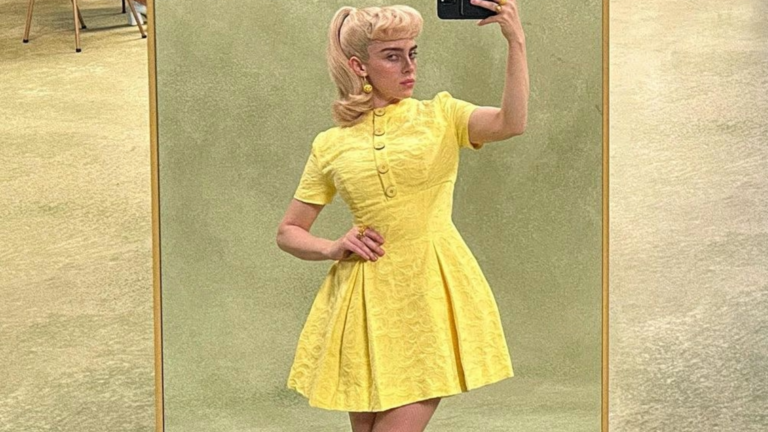 Billie Eilish wearing a yellow dress and a blond wig taking a photo of herself in a mirror against a green backdrop