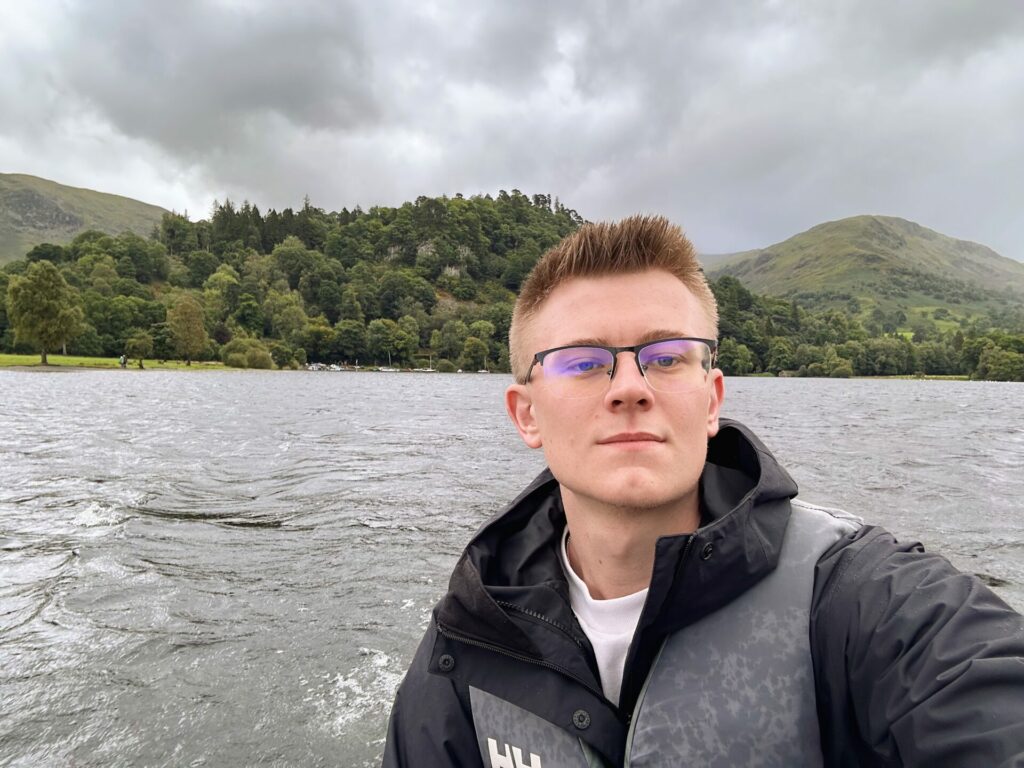 A man looks into the camera with a backdrop of water and tree-lined hills