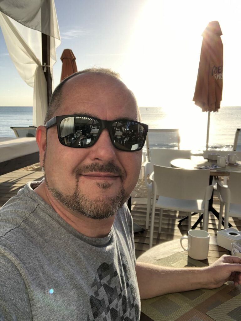 A man wearing a grey T-shirt and tinted sunglasses smiles into the camera