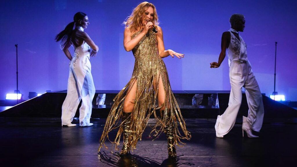 Kylie Minogue performs on stage against a blue backdrop with two dancers