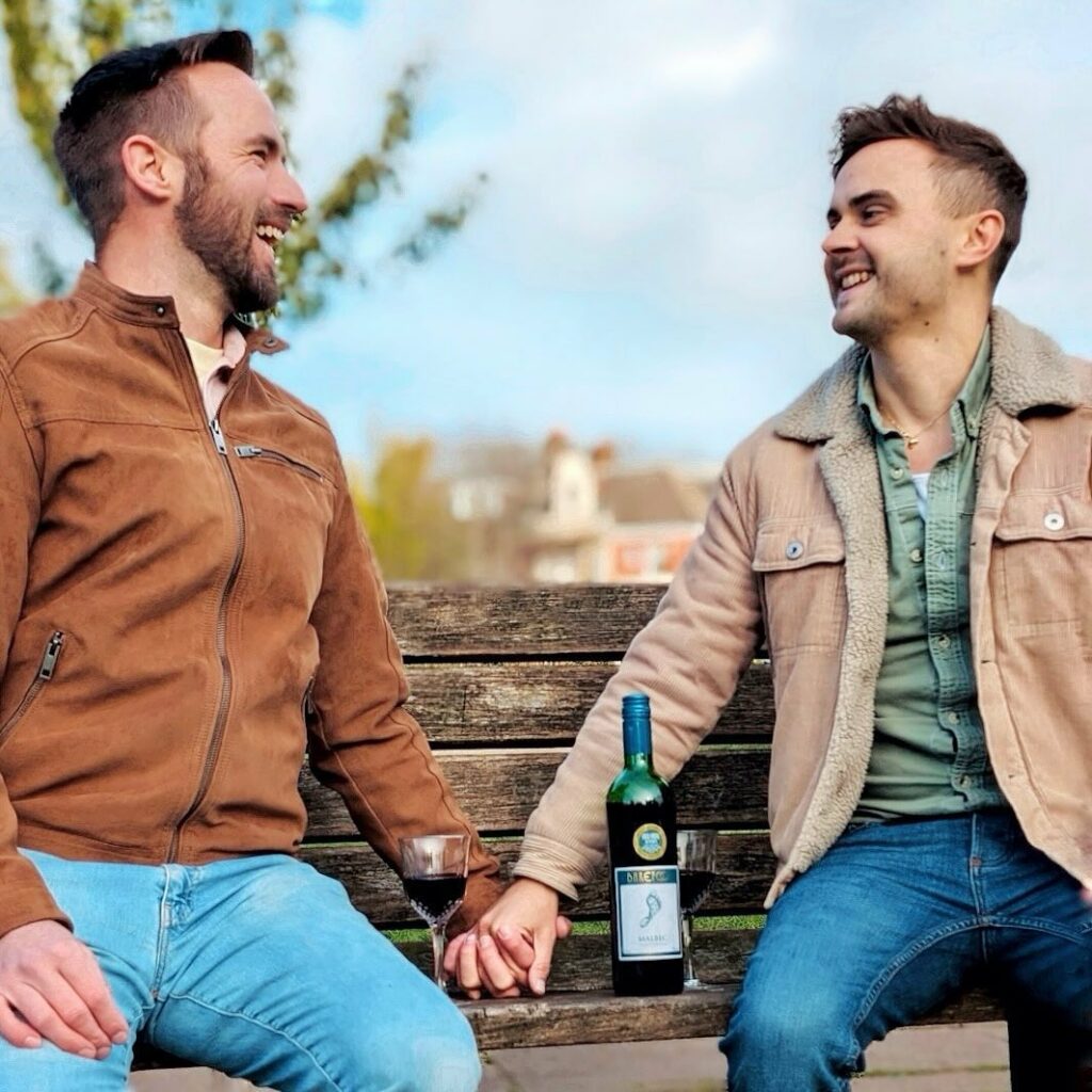 Two smiling men sit on a bench holding hands outdoors with a bottle of Barefoot Wine on the bench