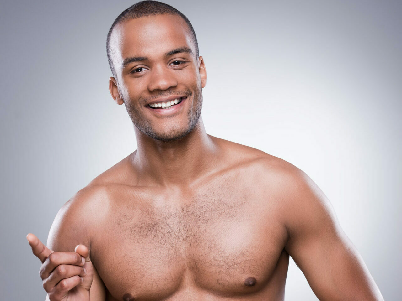 Young shirtless Black man pointing away and smiling while standing against grey background