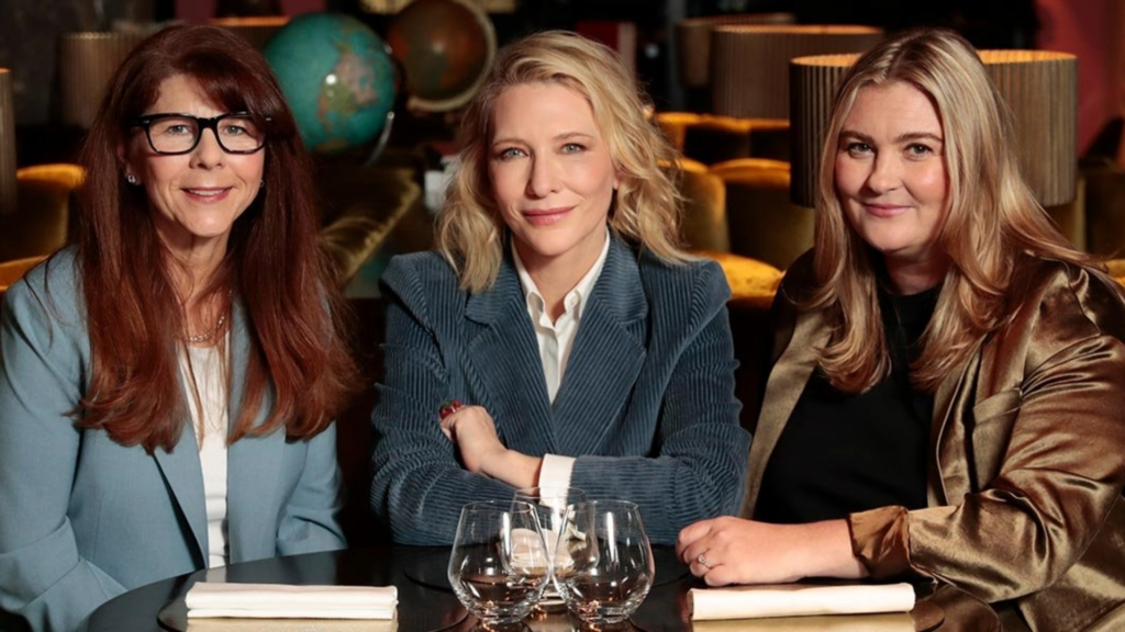 Cate Blanchett sits between two other people at a desk looking into the camera smiling