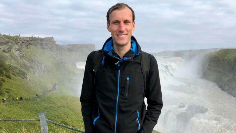 A white man wearing a black coat stands smiling with his hands in his pockets against a background of green misty hills