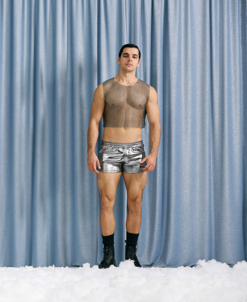 Male model wearing festive clothing poses against a silver curtain backdrop with snow at his feet