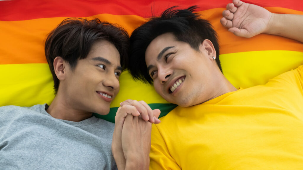 Stock photo of an Asian male same sex couple lying down on a Pride flag looking into each other's eyes and holding hands