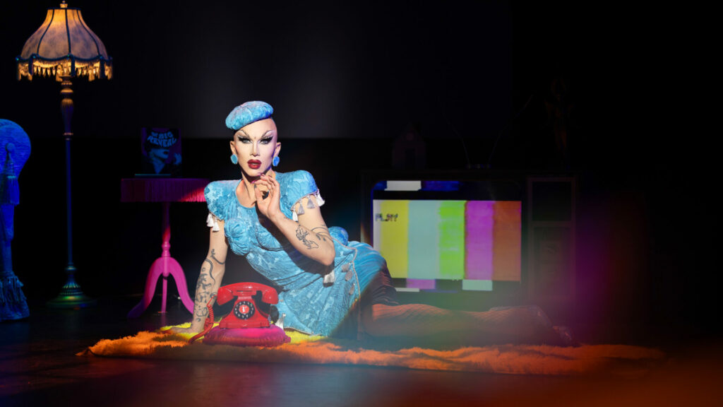 Sasha Velour lies down on a stage in a blue outfit with an old CRT TV behind her and a red telephone in front of her