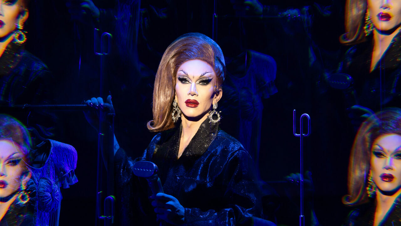 Photo of Sasha Velour onstage in a black outfit surrounded by mirror images of herself