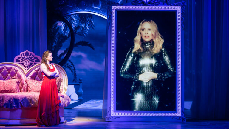 A cast member of I Should Be So Lucky stands onstage next to a large video image of Kylie Minogue