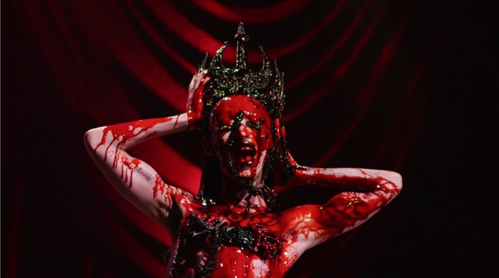 A drag queen wearing a pointed crown is drenched in blood