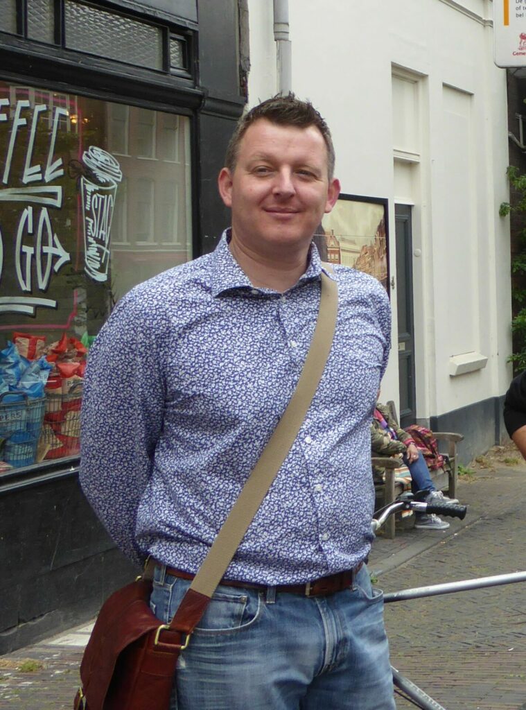 A man wearing a blue shirt stands on the street smiling