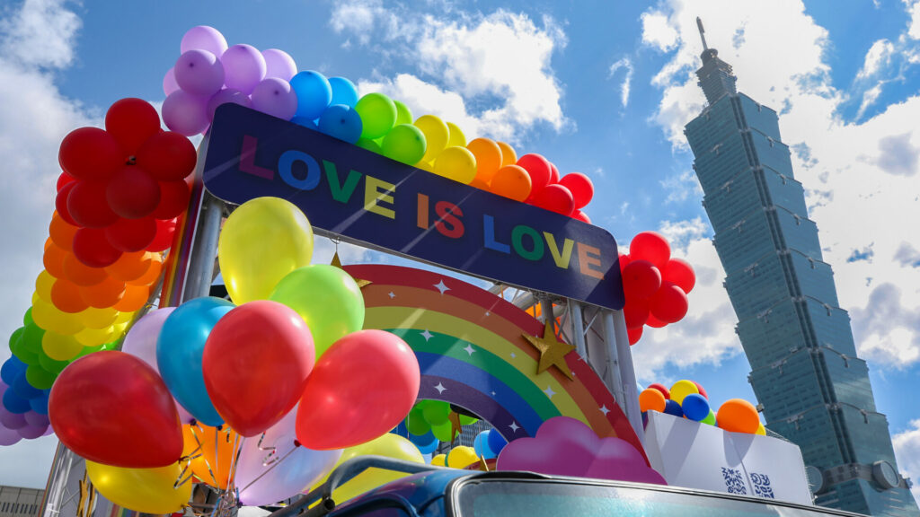 A parade float covered in colourful balloons with a banner sayinjg love is love in front of the Taipei 101 tower and a blue sky