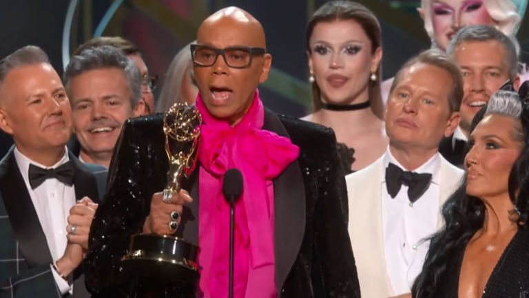 RuPaul stands on stage in a pink shirt and black jacket among castmates from RuPaul's Drag Race holding a golden Emmy award in his hand at the 2024 Emmy Awards