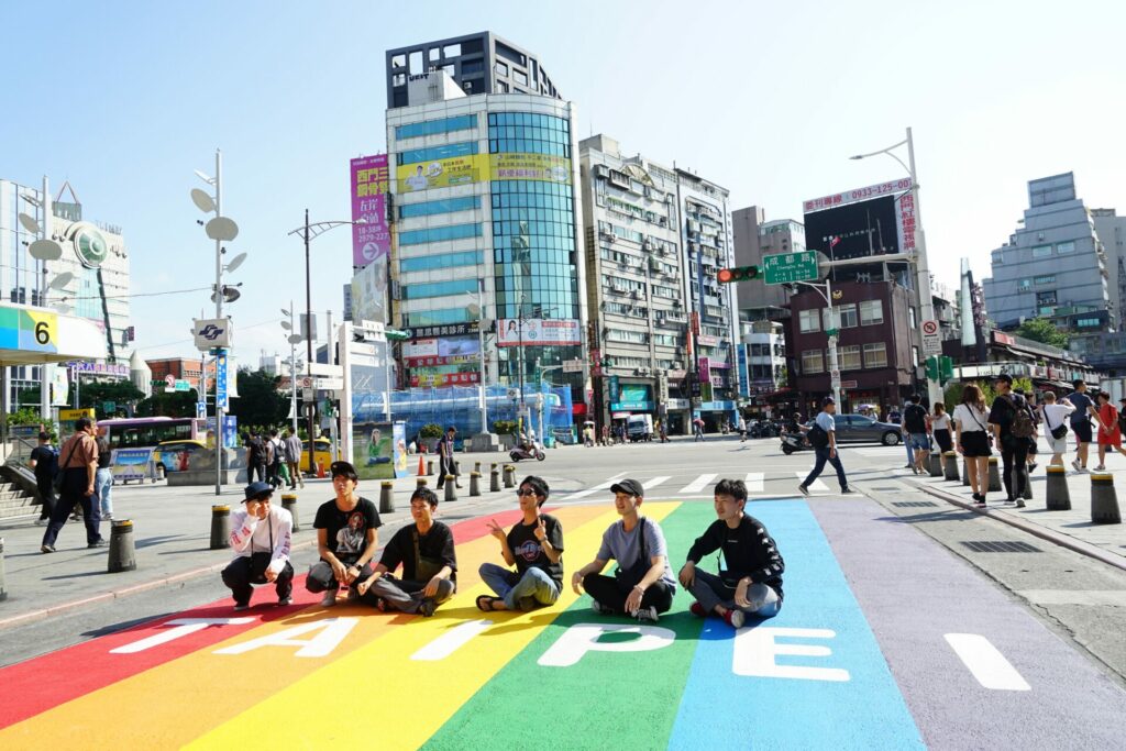 A group of people sit on the ground on a rainbow coloured street crossing with the word TAIPEI on it to pose for a photo