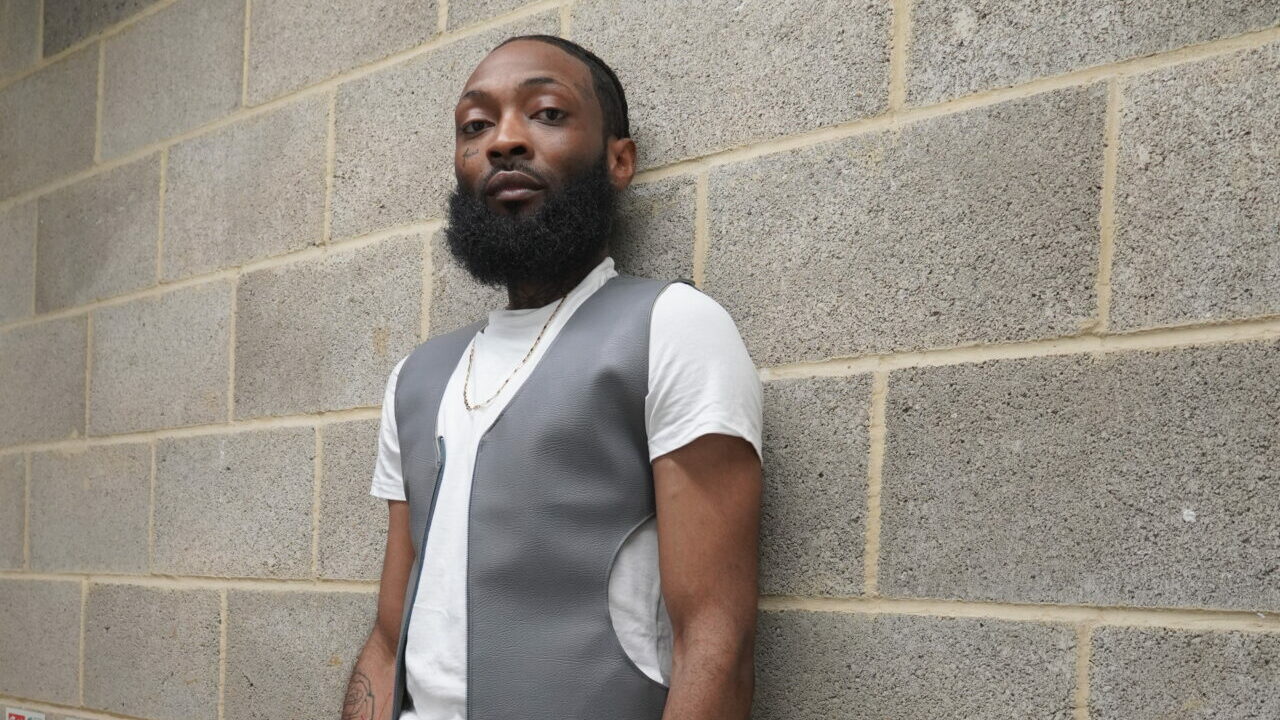 Carter the Bandit stands against a concrete wall wearing a grey waistjacket and white T-shirt