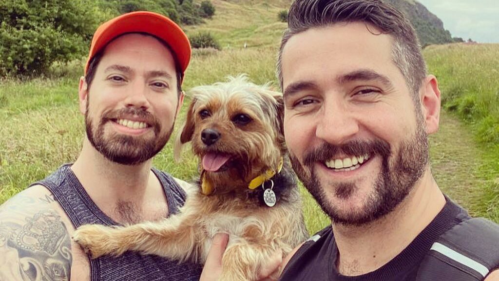 Two smiling men and a dog stand against a backdrop of grass