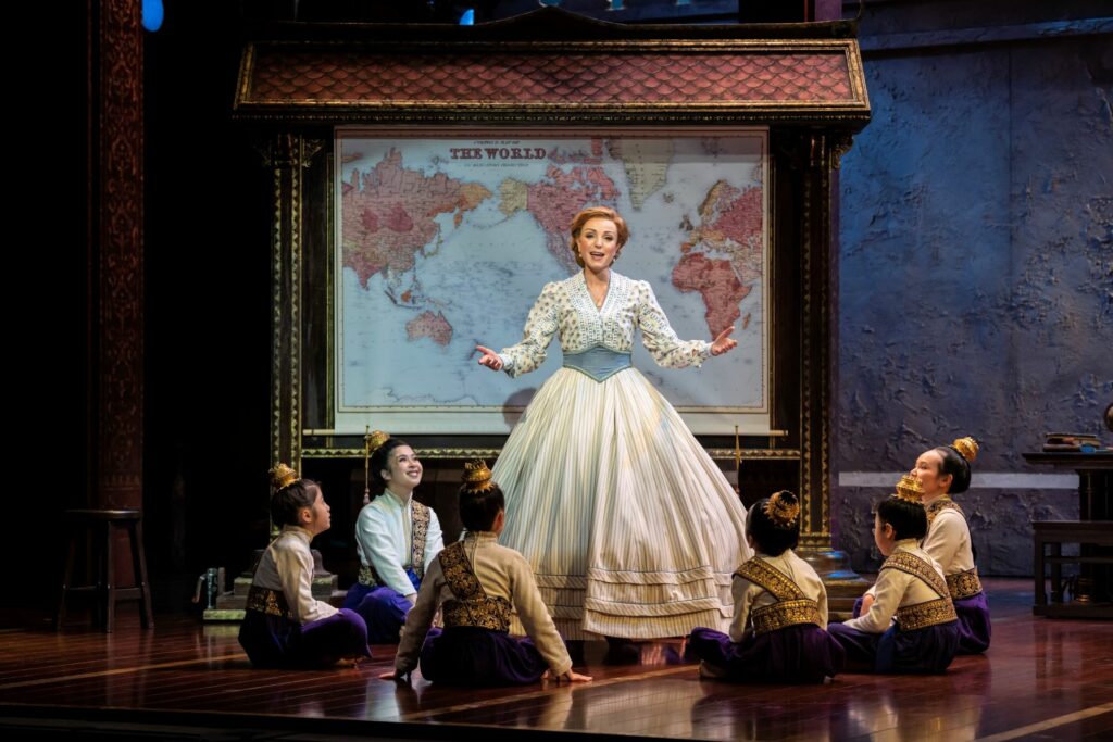 Helen George sings onstage to a group of girls sitting on the floor around her in The King and I