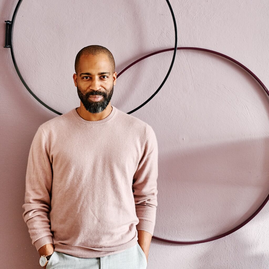 A man wearing a pink jumper stands in front of a pink wall