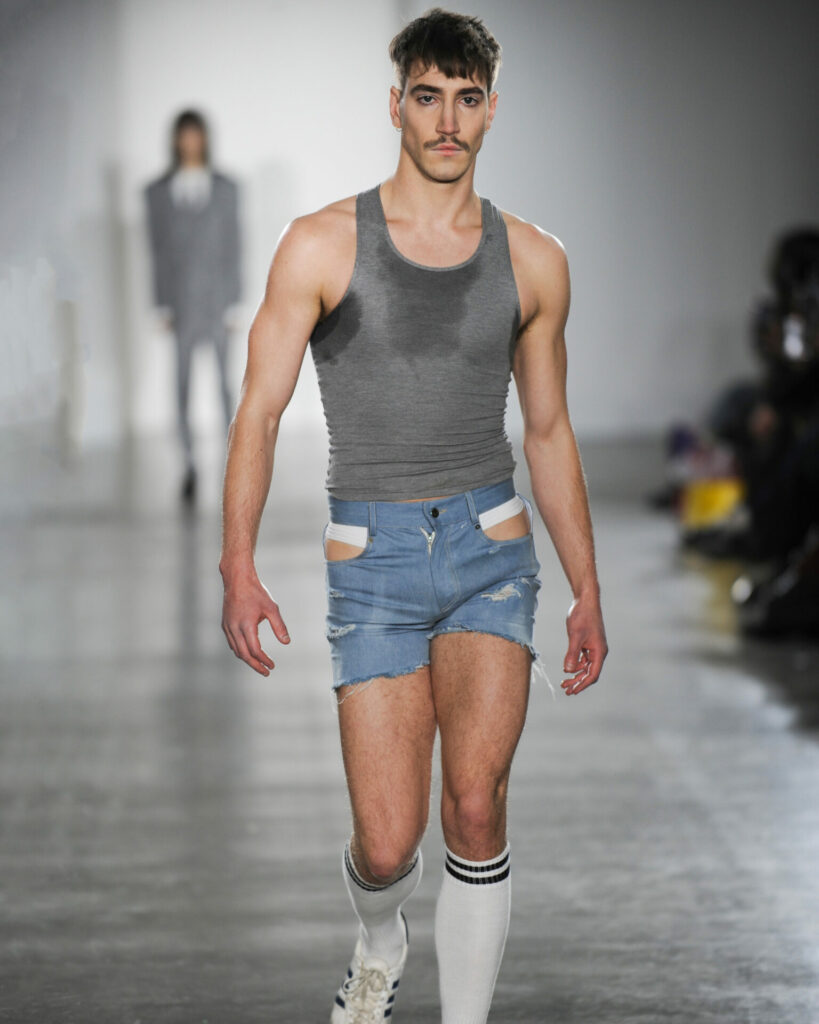 A model walks on a runway wearing denim shorts and a sweaty grey vest