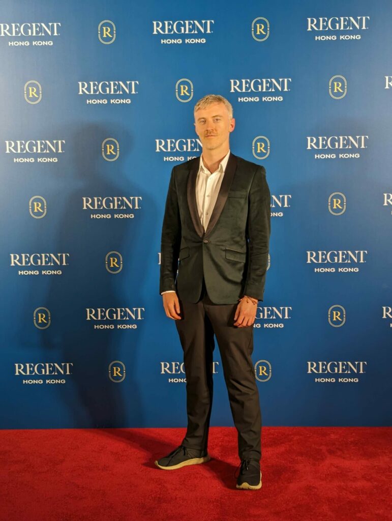 A man stands on a red carpet wearing a black suit