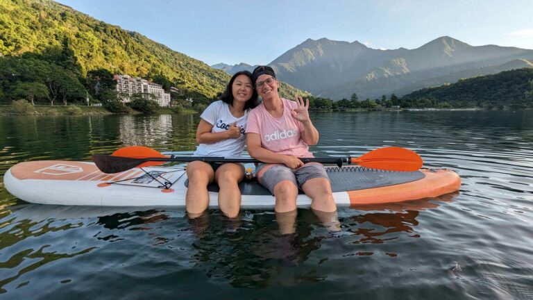Two woman sit on a paddling board together in water with mountains behind them