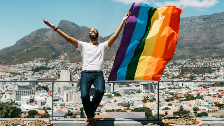 A man stands in front of a cityscape waving a Pride flag