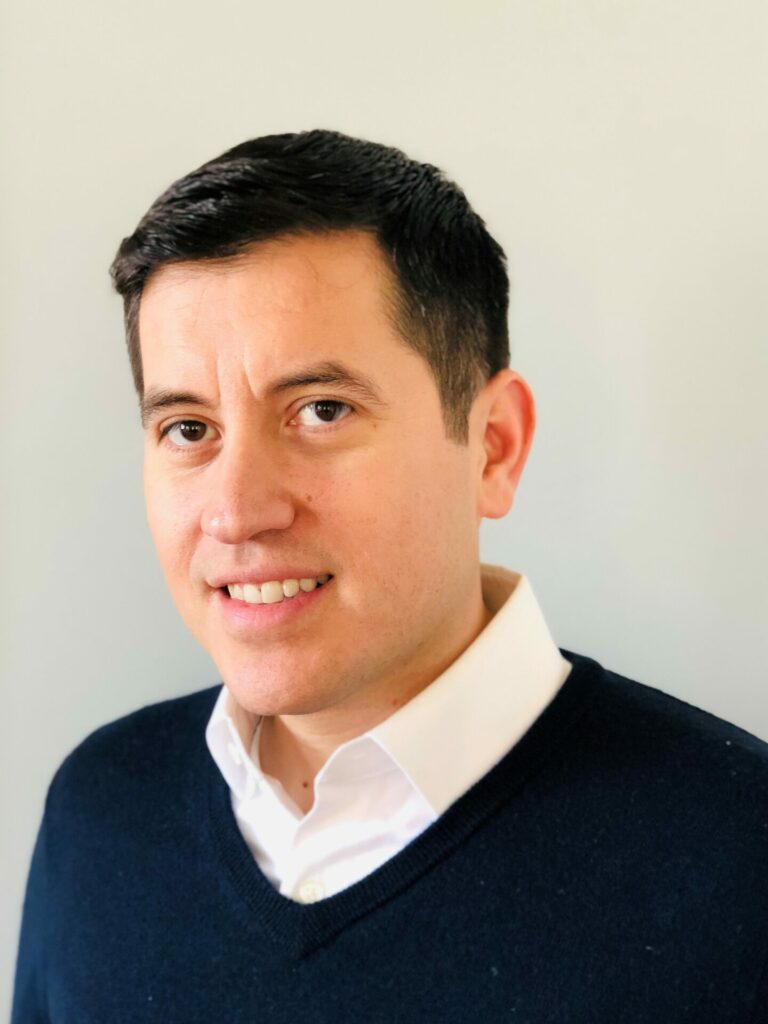 Headshot of a smiling man with a white shirt and blue jumper