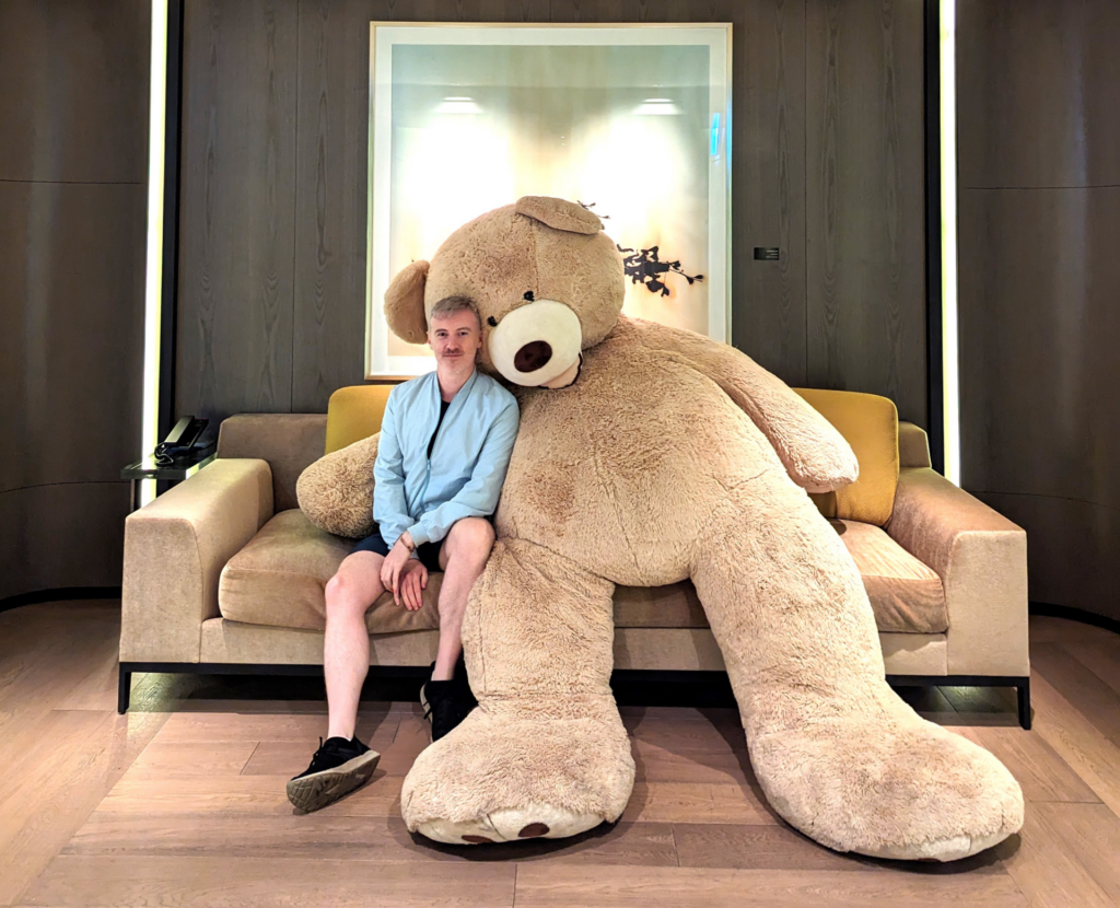 A man sits on a sofa next a giant teddy bear