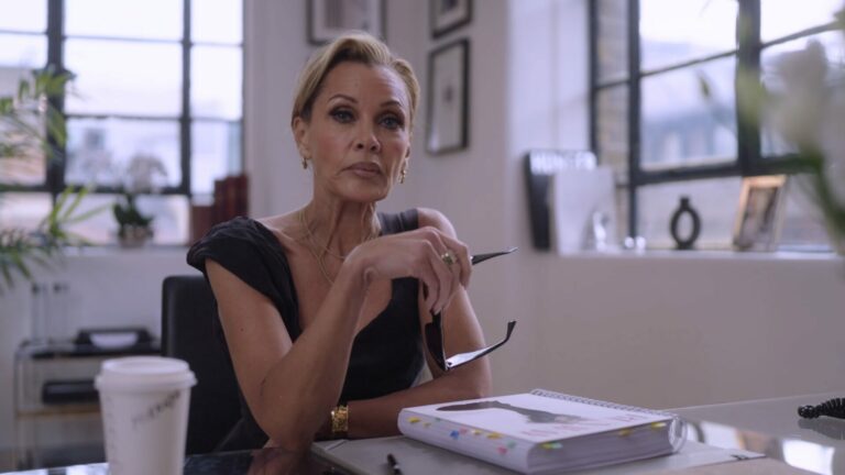 Vanessa Williams sits at a desk holding a pair of sunglasses