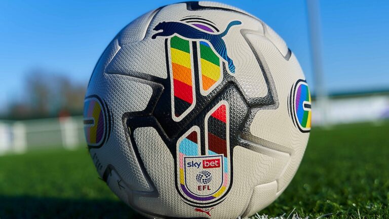 A football with pride colours on it sits on a grass pitch