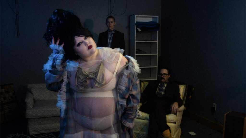 Promo shot of Gossip, featuring Beth Ditto wearing white lingerie with the other two members of the group behind her 