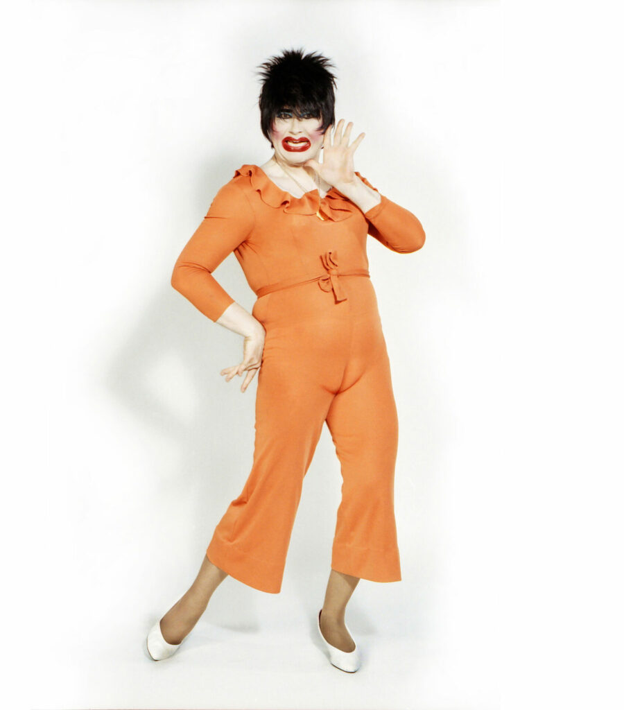A drag queen in an orange jumpsuit with black hair and white shoes