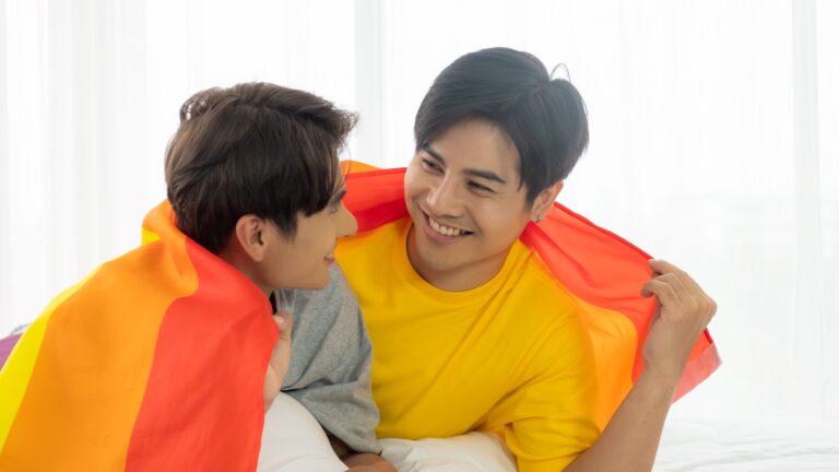 A male couple smile while hugging each other wrapped with a rainbow flag