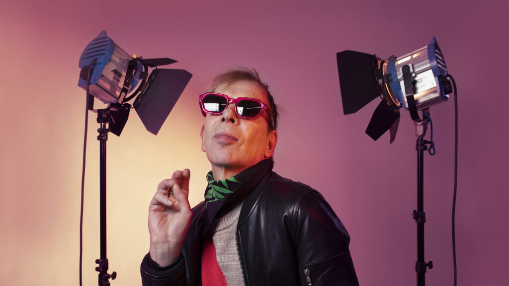 David Hoyle wearing red glasses against a pink backdrop