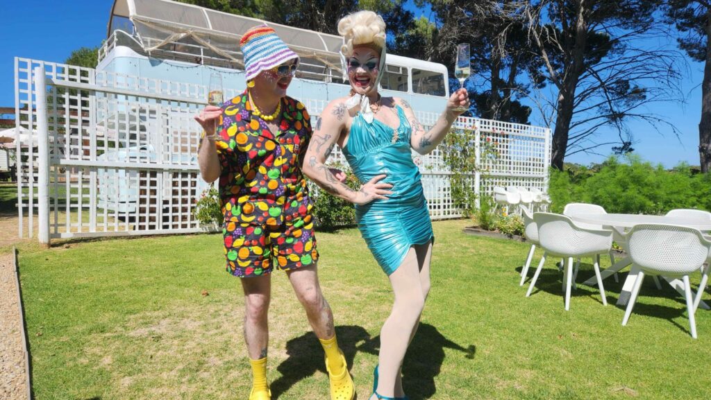 Two drag queens drink wine on a green lawn
