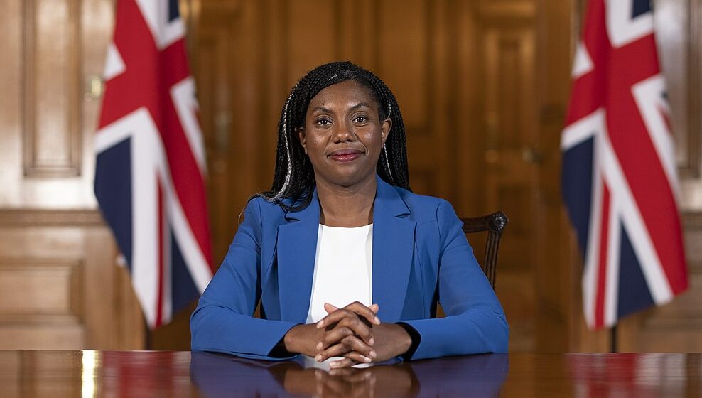 Kemi Badenoch sat at a table with Union Jack flags behind her