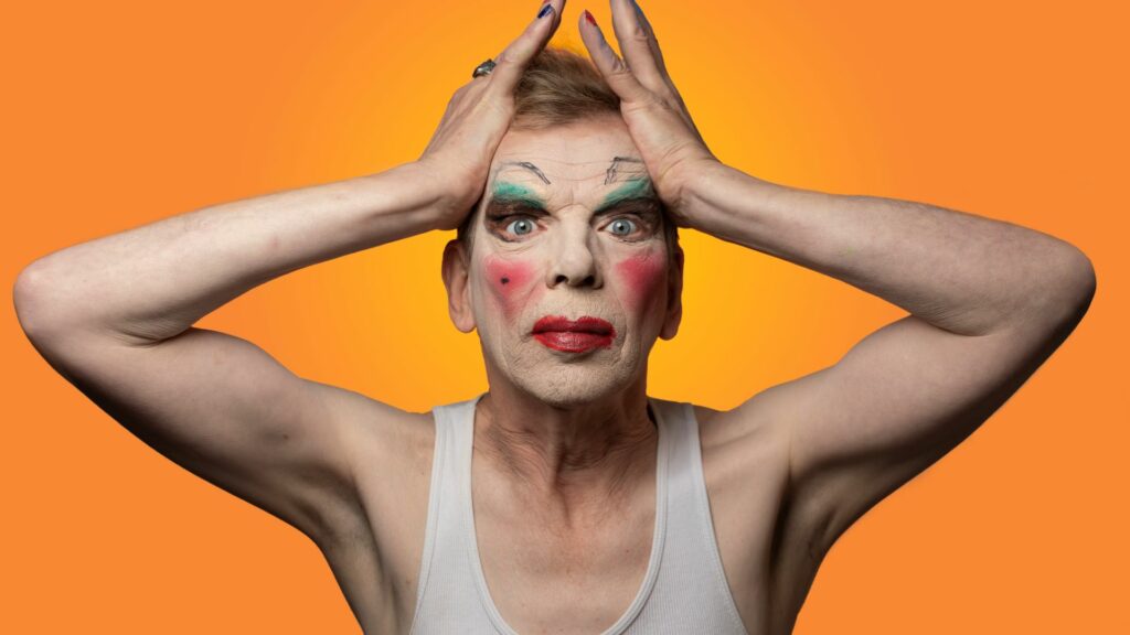 David Hoyle holding his hands on his hand wearing a white vest against an orange backdrop
