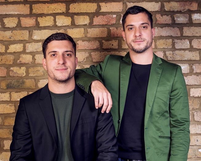 Two twin men wearing suits standing against a brick wall