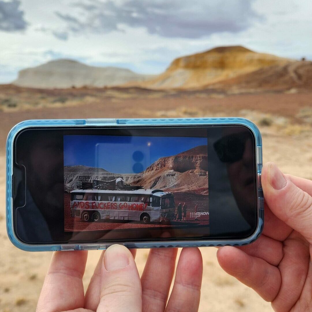 A smartphone being held showing a scene from Priscilla against the actual location