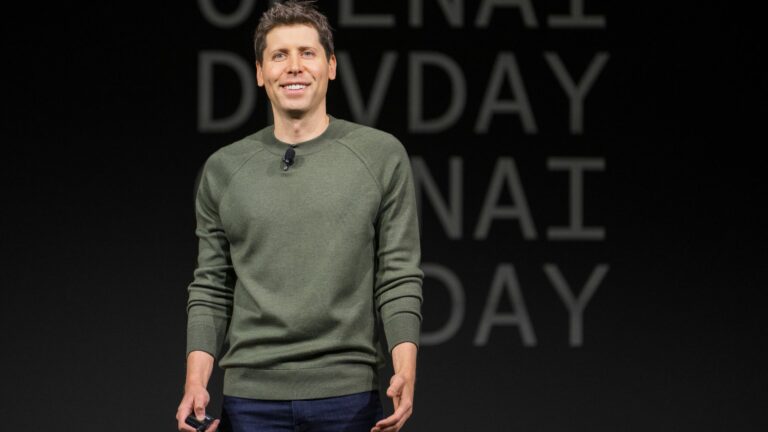 Sam Altman stands on stage smiling wearing a green jumper