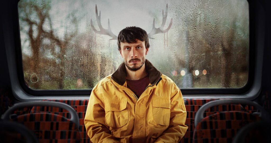 Still from Baby Reindeer showing Richard Gadd sitting on a bus wearing a yellow jacket with a drawing of antlers behind his head