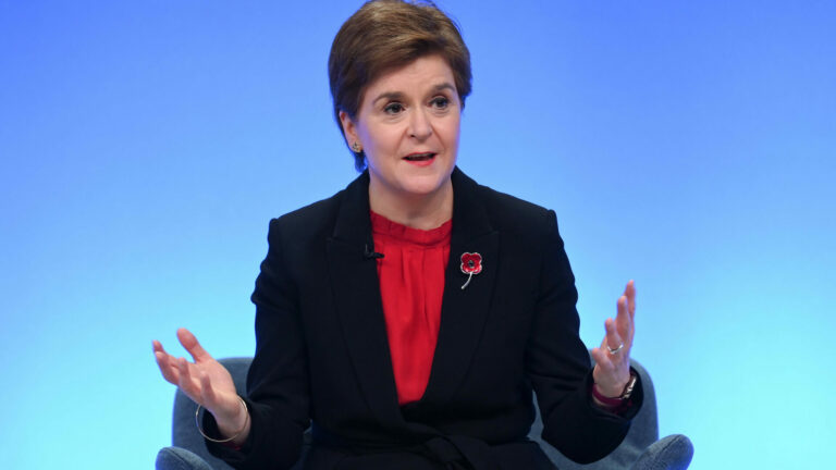 Nicola Sturgeon wearing a black suit and red top against a blue background