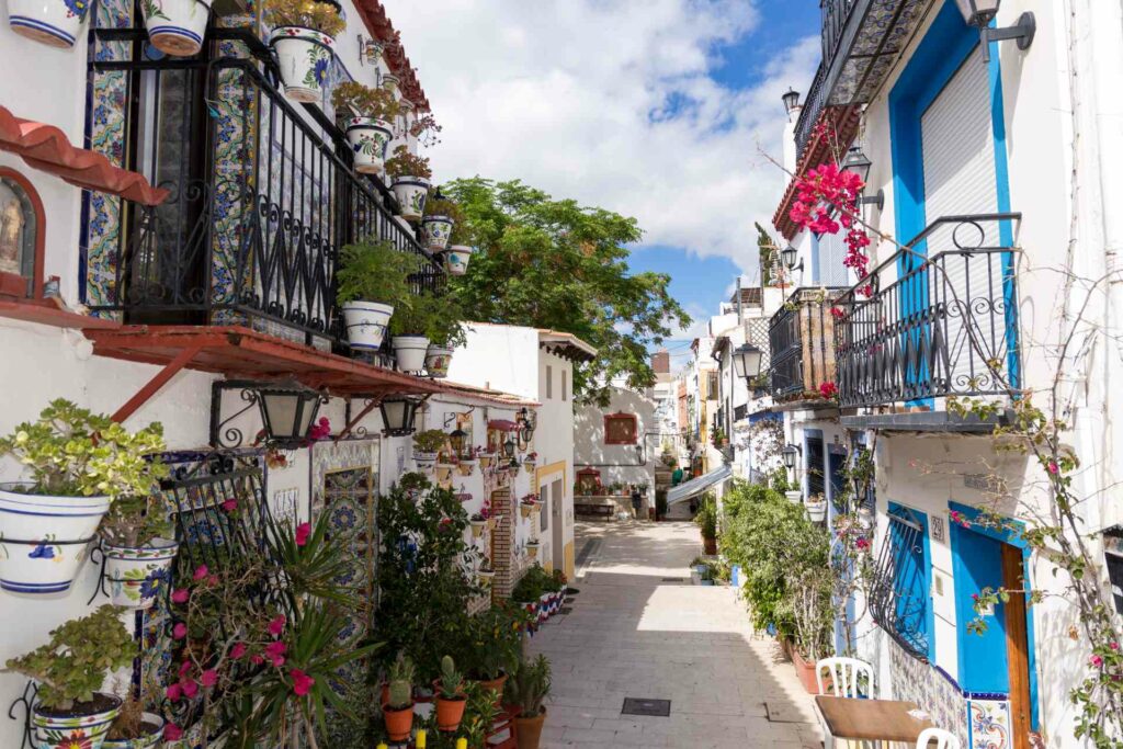 A street in Barrio Santa Cruz, Seville, lined with whitewashed buildings, colourful flowers, and wrought-iron balconies under a bright blue sky.