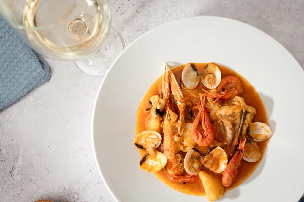 A plate of seafood stew with prawns, clams, and fish in a rich tomato broth, served with a glass of white wine.