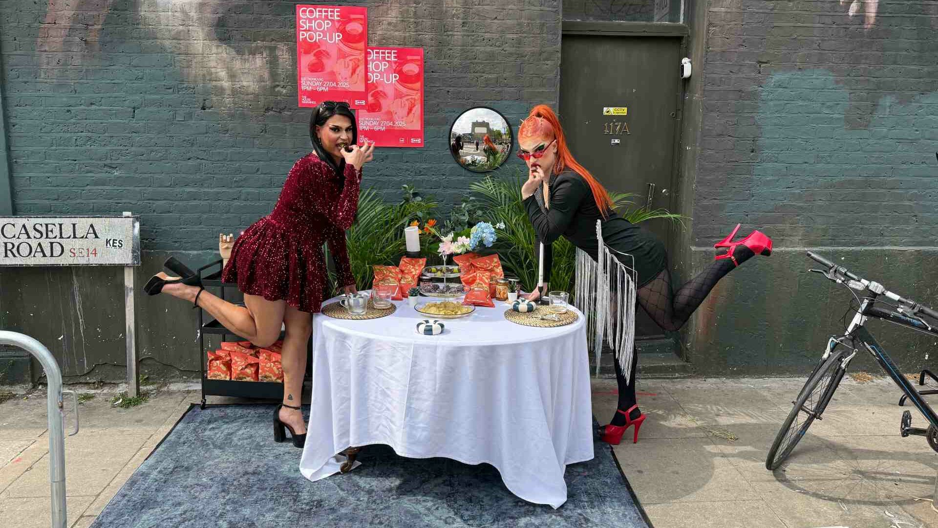 Two drag queens pose playfully beside an outdoor table set with drinks and flowers, in front of a mural-covered wall.