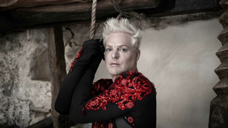 Andy Bell wearing a red top holding on to a rope and looking into the camera