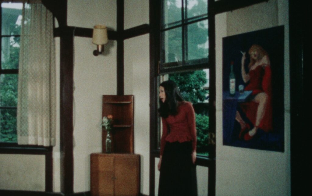 a still from bye bye love, a woman standing in a room in a red jumper